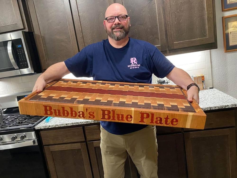 Bubba's Blue Plate owner holding an end grain butcher block with a red center stripe and the name inlaid in red hardwood along the side