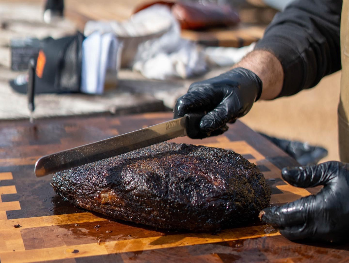 Pitmaster in black gloves slicing a smoked brisket on a walnut end grain butcher block with a staggered hard maple border