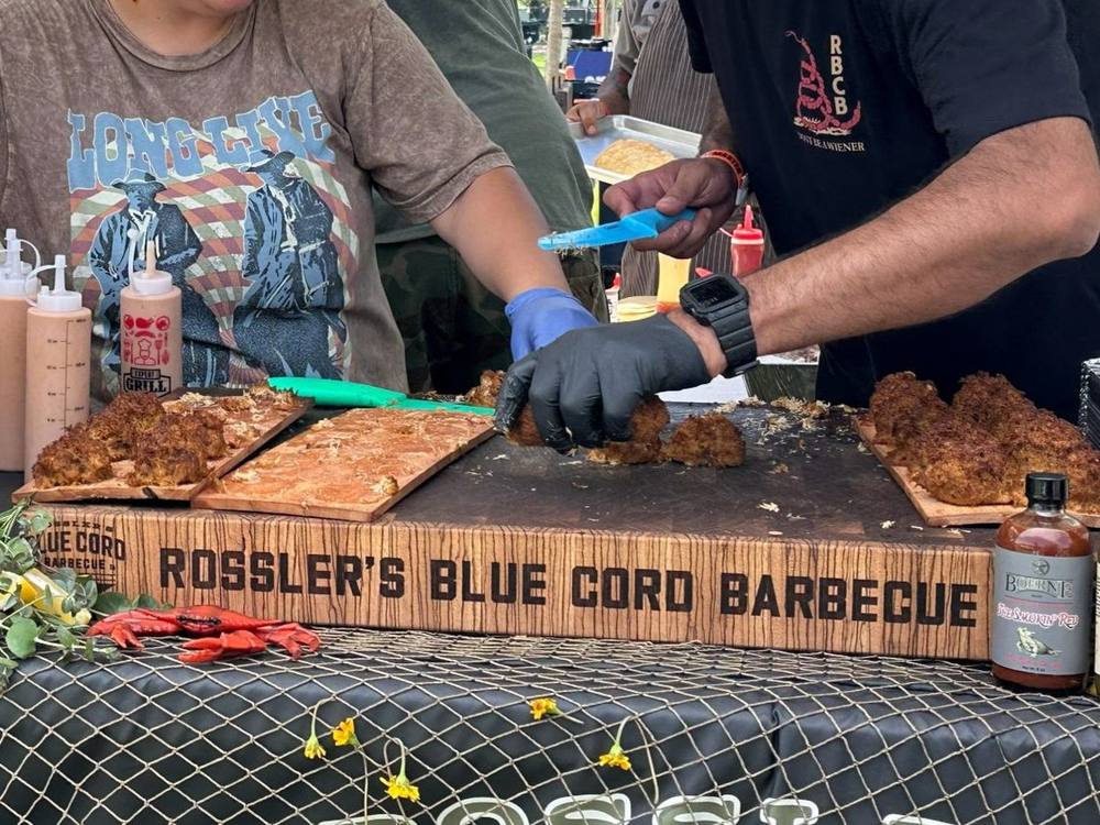Rossler's Blue Cord Barbecue team serving at a festival on their end grain butcher block, with the name along the side